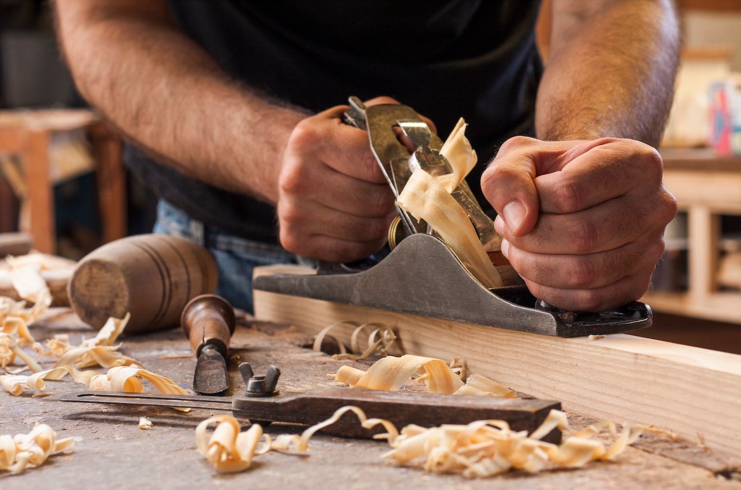 Carpenter hand-planing wood at a workbench with shavings curling off